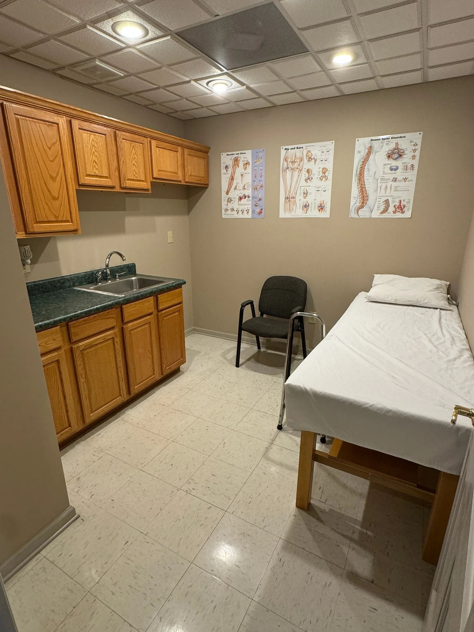 Orthopedic treatment room with an exam table, wooden cabinetry, a sink, and anatomical spine and joint posters on the wall