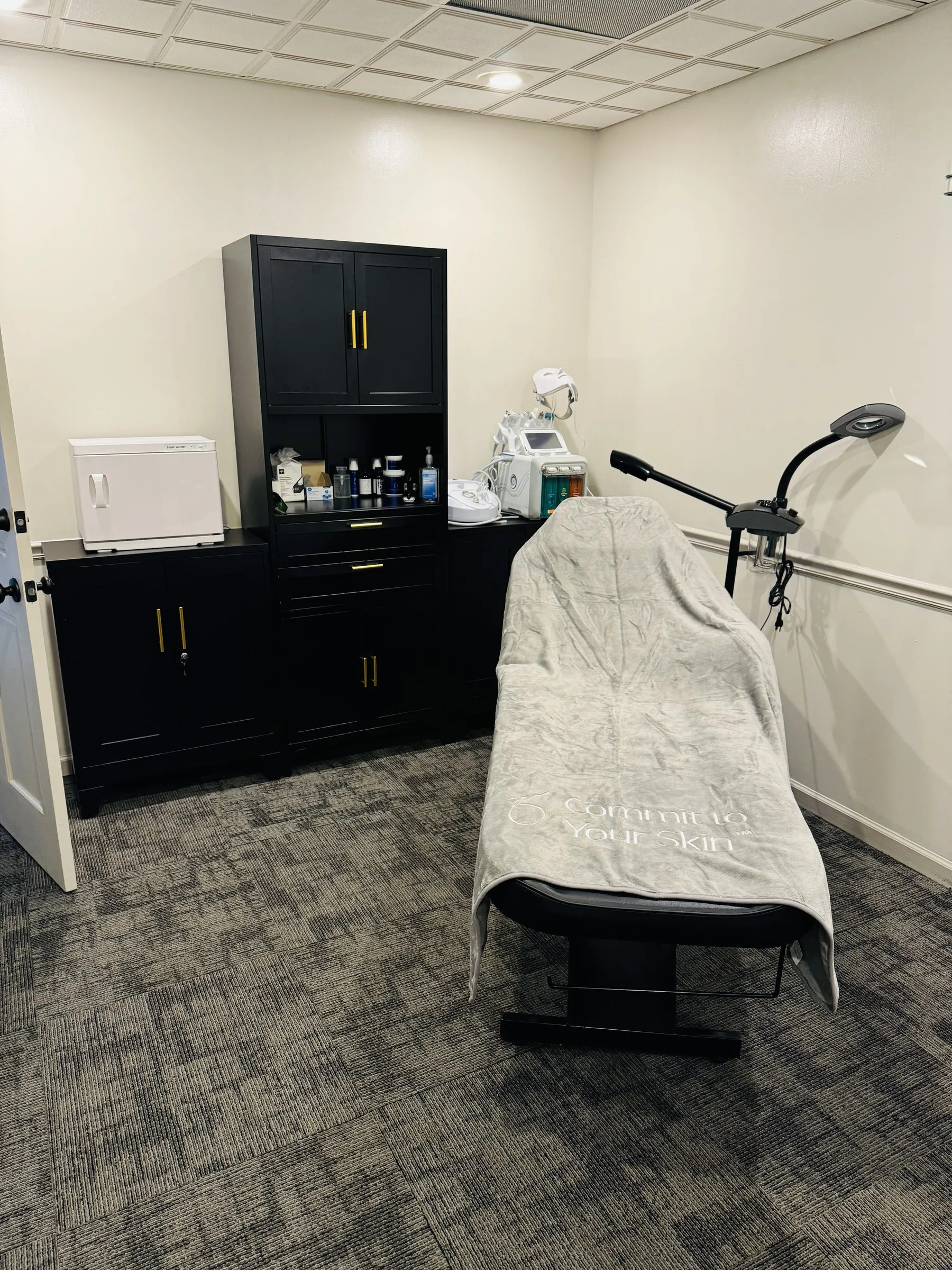 Aesthetics treatment room with a draped treatment table, black cabinetry stocked with skincare products, and skin-care devices